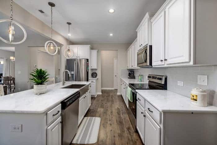 A modern kitchen features white cabinetry, stainless steel appliances, wood flooring, pendant lighting, and a white kitchen island with a sink and dishwasher.