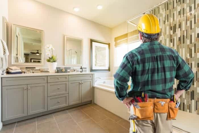 A construction worker wearing a tool belt and yellow hard hat stands in a modern bathroom, facing a vanity and bathtub.