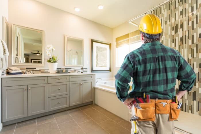 A worker in a yellow hard hat, green plaid shirt, and tool belt stands in a modern bathroom with a large mirror, double vanity, and tiled shower.