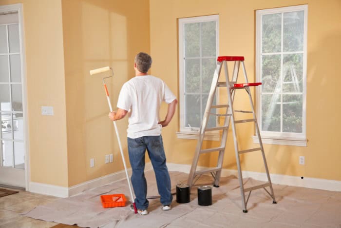 A person standing on a drop cloth, holding a paint roller, looks at a wall next to a step ladder and two paint cans in a room with two windows.