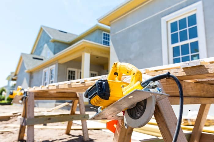 A circular saw rests on a wooden workbench at a residential construction site in front of a partially finished house.