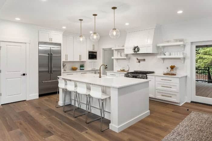 Modern kitchen with white cabinetry, stainless steel appliances, a large island with a white countertop, four white bar stools, and pendant lighting above. Wooden flooring and open shelving are present.